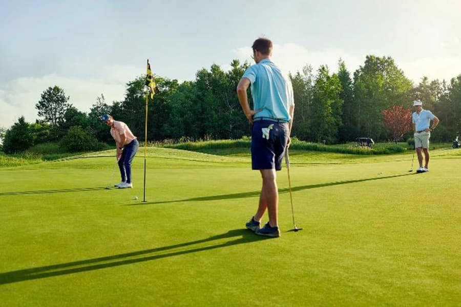 Three golfers are on a green, one is putting while the others watch.