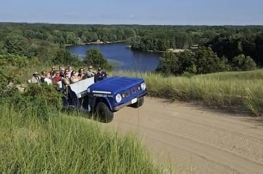 A blue off-road vehicle carries a group of people along a sandy path near a river surrounded by greenery.