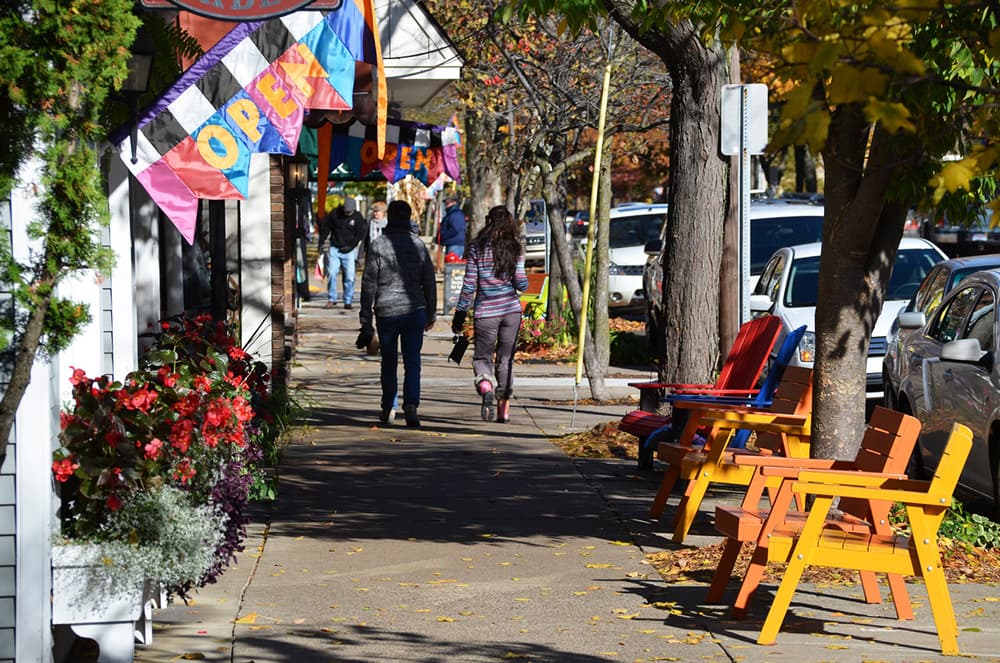 Two people walk along a vibrant, tree-lined sidewalk with colorful chairs and open signs in view.